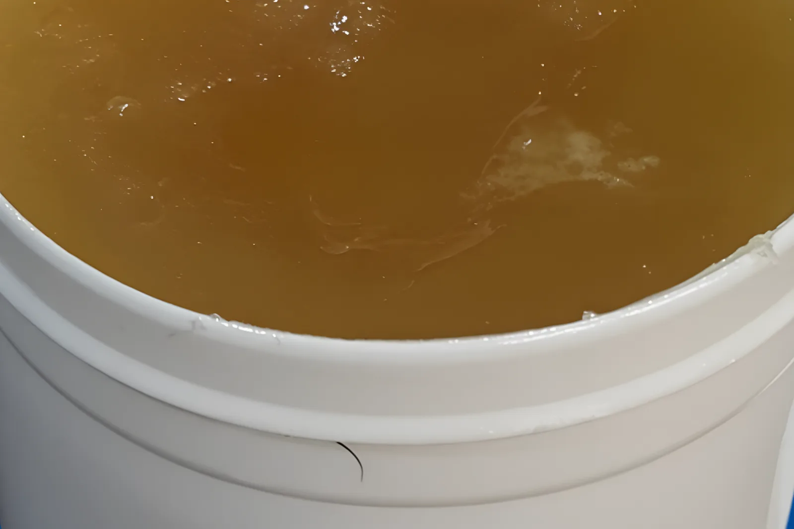Residue wax (foots oil) showing a translucent amber, viscous texture inside a white industrial plastic bucket.